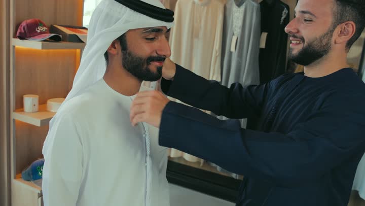 Close-up of a young Emirati man wearing a navy blue kandura taking measurements of a customer's body using a tape measure. Mastering clothing making, men's tailoring and detailing shop, concept of the world of fashion and style, practicing the tailoring profession, skill of mastering detailing youth clothing, handicrafts and crafts.