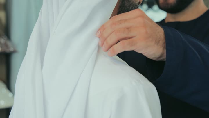 Mastering clothing making, men's tailoring and detailing shop, concept of the world of fashion and style, close-up of a young Emirati man wearing a navy blue kandura taking measurements of a customer's body using a tape measure, practicing the tailoring profession, skill of mastering detailing youth clothing, handicrafts and crafts.