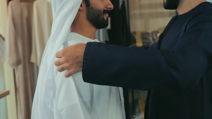 Inventing and creating new and modern models, a men's tailoring and tailoring shop, the concept of the world of fashion and style, a close-up of a young Emirati man wearing a navy blue kandura taking measurements of a customer's body using a tape measure, practicing the tailoring profession, the skill of mastering tailoring youth clothing, handicrafts and crafts.