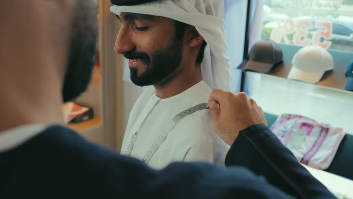 Choosing the best fabrics for tailoring a thobe, a men's tailoring and tailoring shop, the concept of the world of fashion and style, a close-up of a young Emirati man wearing a navy blue kandura taking measurements of a customer's body using a tape measure, practicing the tailoring profession, the skill of mastering tailoring youth clothes, handicrafts and crafts.