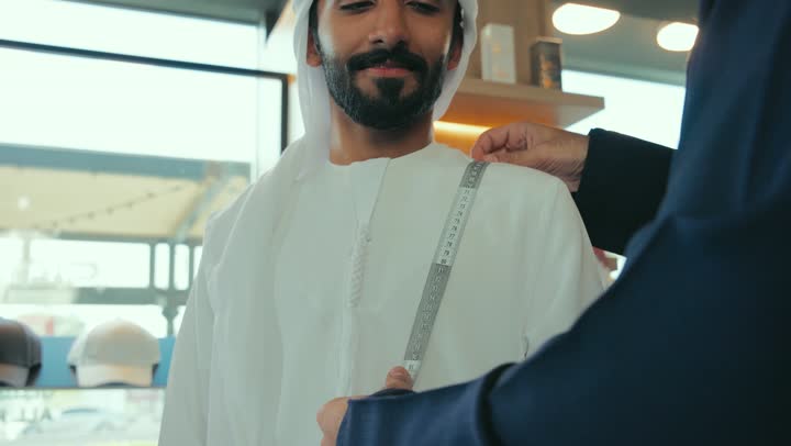 محل خياطة و تفصيل الملابس الرجالي ، مفهوم عالم الازياء والموضه ، تصوير مقرب لشاب خليجي اماراتي يرتدي الكندورة الكحلي يقوم بأخذ القياسات الخاصة بجسم الزبون باستخدام الشريط المتري ، ممارسة مهنة الخياطة، مهارة اتقان تفصيل الملابس الشبابية، حرف ومهن يدوية .