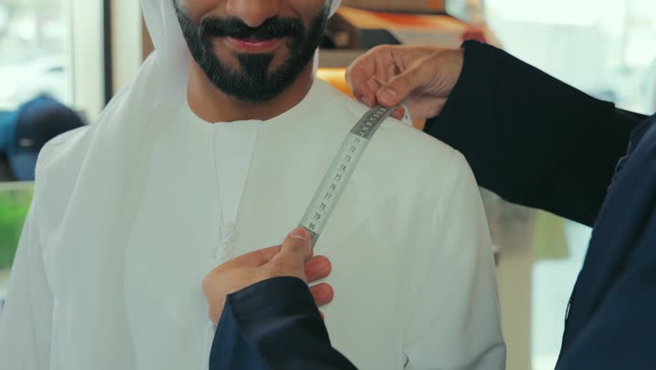 مفهوم عالم الازياء والموضه ،  تصوير مقرب لشاب خليجي اماراتي يرتدي الكندورة الكحلي يقوم بأخذ القياسات الخاصة بجسم الزبون باستخدام الشريط المتري ، ممارسة مهنة الخياطة، مهارة اتقان تفصيل الملابس الشبابية، حرف ومهن يدوية، محل خياطة و تفصيل الملابس الرجالي  .