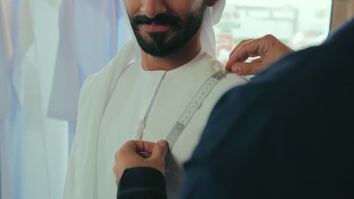 Close-up of a young Emirati man wearing a navy blue kandura taking measurements of a customer's body using a tape measure, practicing the tailoring profession, skill of mastering the tailoring of youth clothes, handicrafts and crafts, a men's tailoring and tailoring shop.