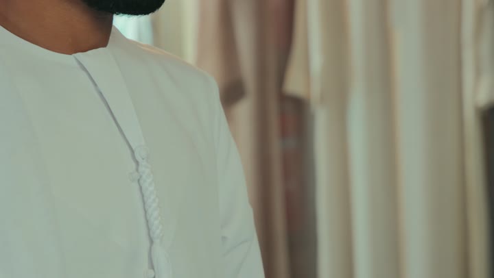 ممارسة مهنة الخياطة، مهارة اتقان تفصيل الملابس الشبابية، حرف ومهن يدوية، محل خياطة و تفصيل الملابس الرجالي ،  تصوير مقرب لشاب خليجي اماراتي يرتدي الكندورة الكحلي يقوم بأخذ القياسات الخاصة بجسم الزبون باستخدام الشريط المتري .