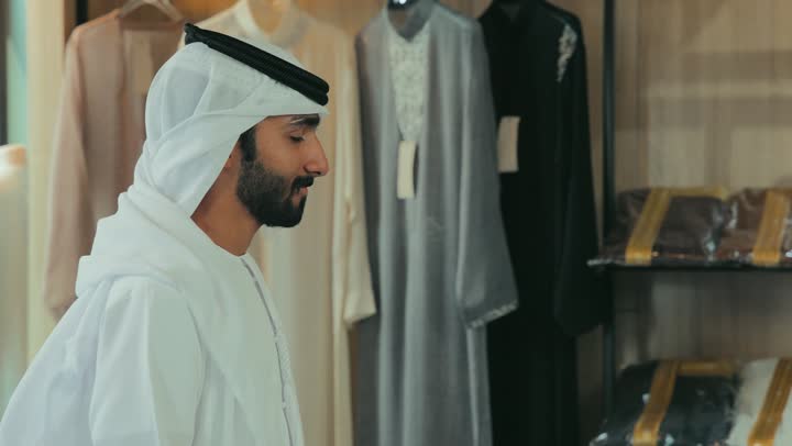مهارة الخياطة واتقان تفصيل الملابس الشبابية، حرف ومهن يدوية، محل خياطة و تفصيل الملابس الرجالي ، ممارسة مهنة الخياطة، تصوير مقرب لشاب خليجي اماراتي يرتدي الكندورة الكحلي يقوم بأخذ القياسات الخاصة بجسم الزبون باستخدام الشريط المتري .