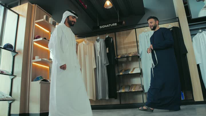 Handicrafts and crafts, men's tailoring and tailoring shop, practicing the tailoring profession, close-up of a young Emirati Gulf man wearing a navy blue kandura taking measurements of a customer's body using a tape measure, sewing skill and mastery of tailoring youth clothing