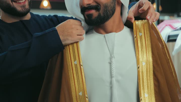 The joy of shopping and buying, a young Emirati man wearing a kandura trying on clothes, a picture of a salesman helping a customer put on a brown Emirati bisht with gestures of happiness and pleasure, taking appropriate measurements, the art of sewing and fashion design, a men's clothing store.