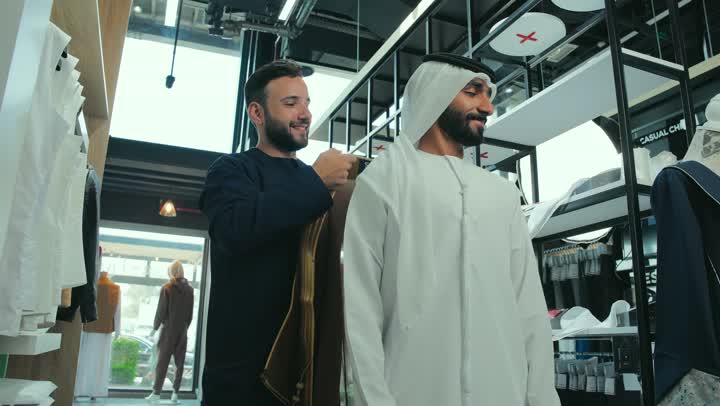 A young Emirati man wearing a kandura trying on clothes, a picture of a salesman helping a customer put on a brown Emirati bisht with gestures of happiness and pleasure, taking appropriate measurements, the art of sewing and fashion design, a men's clothing store, the pleasure of shopping and buying
