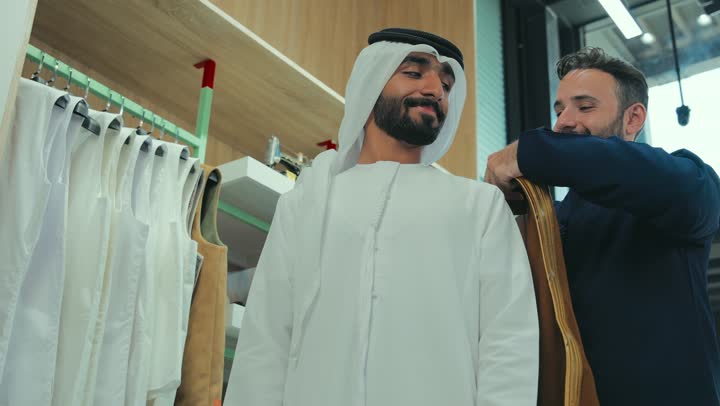 A photo of a salesman helping a customer put on a brown Emirati bisht with gestures of happiness and pleasure, taking appropriate measurements, the art of tailoring and fashion design, a men's clothing store, a young Emirati Gulf man wearing a kandura trying on clothes, the joy of shopping and buying