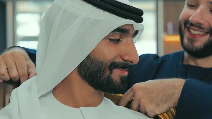 Taking the right measurements, the art of tailoring and fashion design, a men's clothing store, a picture of a salesman helping a customer put on a brown Emirati bisht with gestures of happiness and pleasure, a young Emirati Gulf man wearing a kandura trying on clothes, the joy of shopping and buying