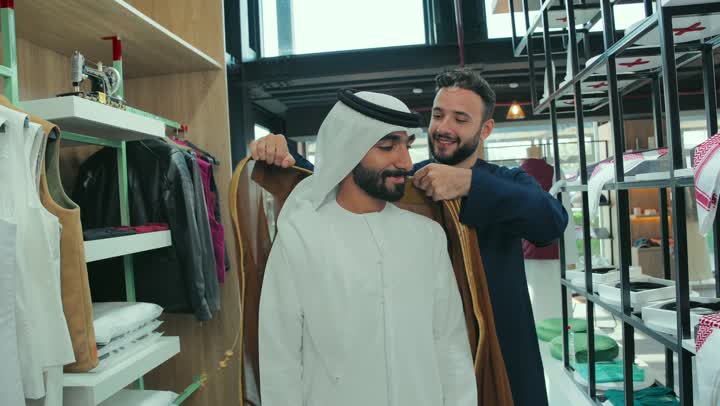 فن الخياطة وتصميم ازياء، محل تفصيل الملابس الرجالي، أخذ القياسات المناسبة ، تصوير لبائع يقوم بمساعدة الزبون بارتداء بشت اماراتي  بني اللون بايماءات السعاده و السرور، شاب خليجي اماراتي يرتدي الكندورة يقوم بقياس الملابس ، متعه التسوق و الشراء