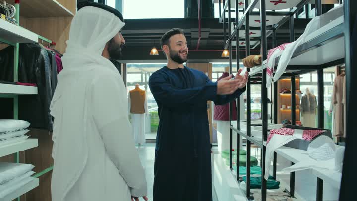 Men's tailoring and detailing shop in the Emirates, the concept of the world of fashion and style, the selection of the finest fabrics, a close-up shot of the hand of a young Emirati Gulf Arab man wearing a white kandura inside a shop specializing in tailoring men's clothing, the skill of sewing and mastery of detailing youth clothing.