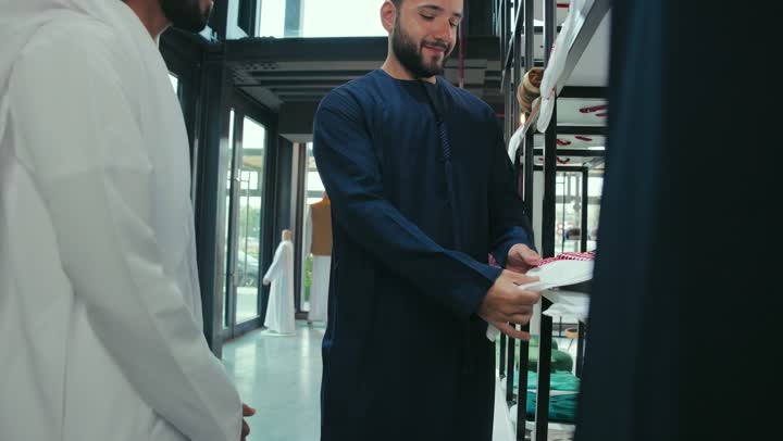 A close-up of a young Emirati Gulf Arab man wearing a navy blue kandura, working as a tailor, showing customers the finest fabrics for tailoring thobes. Mastery of tailoring clothes, enjoying shopping and buying clothes, a men's tailoring and tailoring shop.