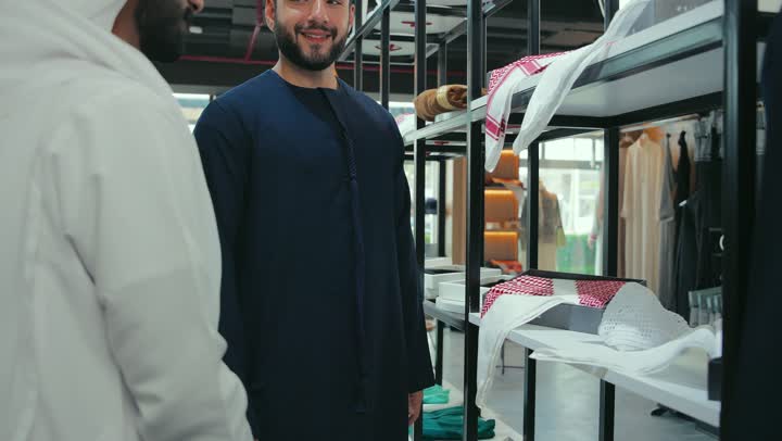 Men's tailoring and tailoring shop, enjoy shopping and buying clothes, mastery of tailoring clothes, close-up photo of a young Emirati Gulf Arab man wearing a navy blue kandura working in tailoring and showing the customer the finest types of fabrics for tailoring the thobe