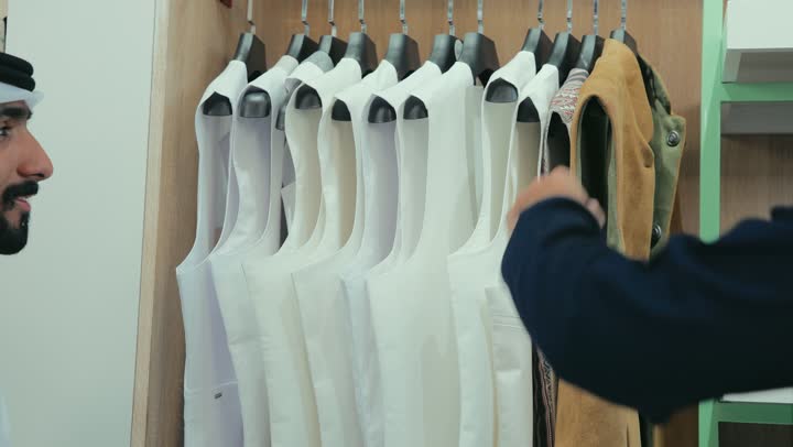 Choosing the finest fabrics for making clothes, a men's tailoring and tailoring shop, the pleasure of shopping and buying clothes, a photo of a young Emirati Gulf Arab man wearing a navy blue kandura working as a tailor, displaying a winter jacket to a customer