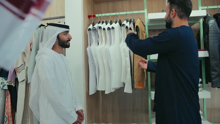 Men's tailoring and detailing shop, choosing the finest fabrics for making clothes, the pleasure of shopping and buying clothes, a photo of a young Emirati Gulf Arab man wearing a navy blue kandura working as a tailor, displaying a winter jacket to a customer