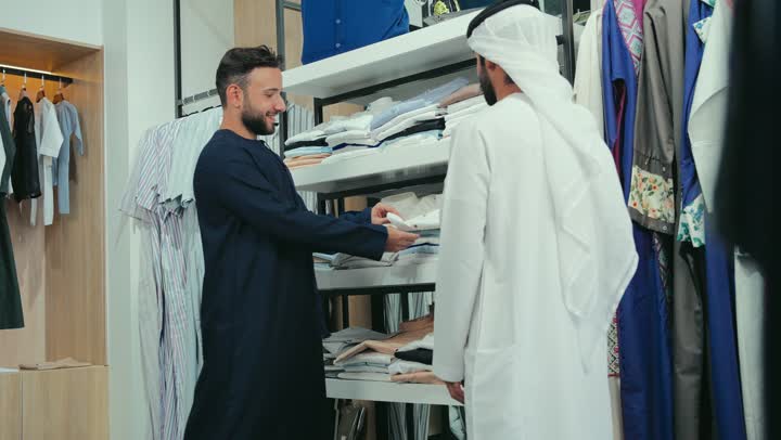 مفهوم عالم الأزياء والموضة، اقتناء ملابس جديدة، خدمه العملاء في المتاجر، مهارة الخياطة واتقان تفصيل الملابس الشبابية، تصوير لشاب عربي خليجي اماراتي متبسم يرتدي الكندورة الكحلي يعمل في مهنة الخياطة يقوم بعرض قميص ابيض للزبون