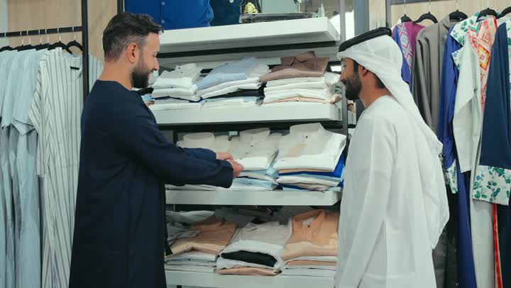 خدمه العملاء في المتاجر، مفهوم عالم الأزياء والموضة، مهارة الخياطة واتقان تفصيل الملابس الشبابية، اقتناء ملابس جديدة، تصوير لشاب عربي خليجي اماراتي متبسم يرتدي الكندورة الكحلي يعمل في مهنة الخياطة يقوم بعرض قميص ابيض للزبون