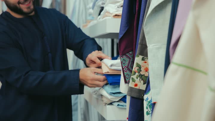 Gestures of enjoying shopping, the concept of the world of fashion, buying men's supplies from the market, a photograph of the hand of a smiling Emirati Gulf Arab customer wearing a navy blue kandura choosing clothes from an Emirati store.