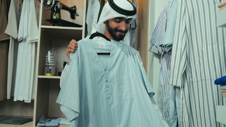 شراء القمصان الجديده ، تصوير لرجل عربي خليجي اماراتي يرتدي الكندوره والغتره البيضاء داخل المتجر يقوم باختيار قميص، قضاء وقت ممتع في شراء الملابس الرجالية، الاستمتاع بالتسوق والاستفاده من العرض و الخصم الموسمي.