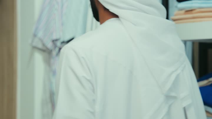A photo of an Emirati Gulf Arab man wearing a white kandura and ghutra inside a store choosing a shirt, having a good time shopping for men's clothing, enjoying shopping and taking advantage of the seasonal offer and discount.