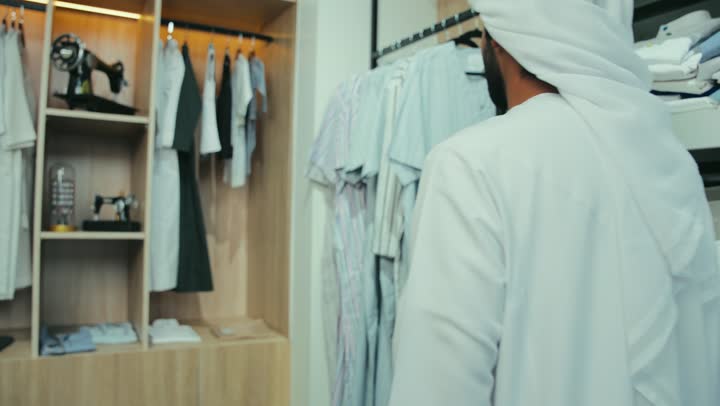 Having a great time shopping for men's clothing. A picture of an Emirati Gulf Arab man wearing a white kandura and ghutra inside a store choosing a shirt. Enjoy shopping and take advantage of the seasonal offer and discount.