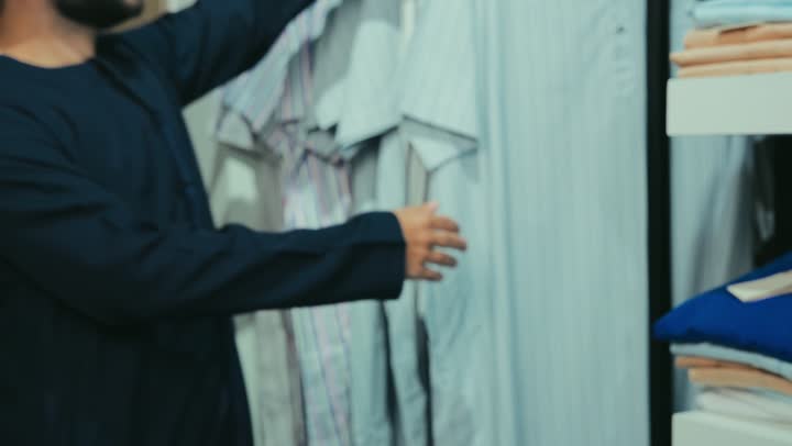 Gestures of happiness and pleasure, taking advantage of the seasonal offer and discount, close-up of a smiling Emirati Gulf Arab customer wearing a navy blue kandura choosing a thobe from a men's accessories store, enjoying shopping in a mall in the United Arab Emirates