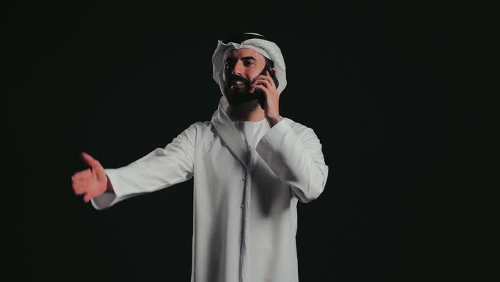 إيماءات وتعابير السعادة والسرور لسماع الأخبار المفرحة، التواصل مع الأهل والأصدقاء، تصوير لرجل عربي خليجي إماراتي مبتسم يرتدي الكندورة والغترة يقوم بإجراء مكالمة هاتفية عبر الهاتف المحمول، استخدام التقنيات والأجهزة الحديثة