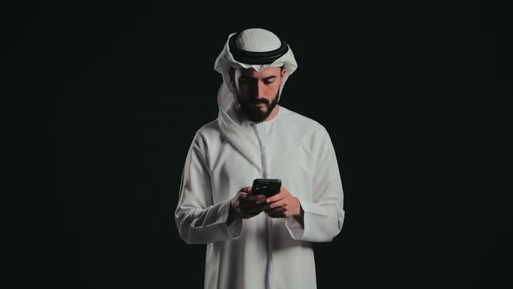 الدردشه عبر الانترنت ، تصفح مواقع التواصل الاجتماعى ، استخدام التكنولوجيا المتطورة للتسويق الرقمي الإلكتروني، عرض تطبيقات و خدمات الهاتف المحمول، تصوير لشاب عربي خليجي اماراتي يرتدي الكندورة والغترة يستخدم جهاز الجوال .