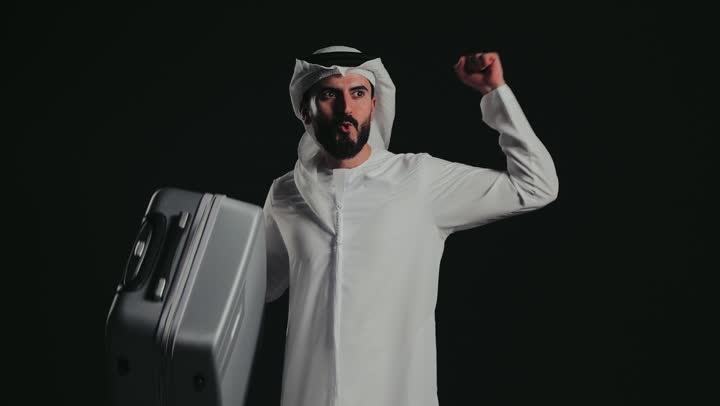 The concept of tourism and travel during the summer vacation, taking a leisure tour, photographing an Emirati Gulf Arab man wearing a kandura and ghutra carrying a luggage bag, looking at the camera with gestures of pleasure, joy and readiness to spend an enjoyable summer trip.