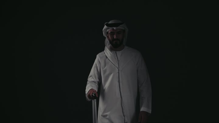 Taking a leisure trip, a picture of an Emirati Gulf Arab man wearing a kandura and ghutra standing next to his luggage, looking at a place with gestures of pleasure, joy and readiness to spend a pleasant summer trip, the concept of tourism and travel during the summer vacation.