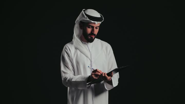 Writing and recording notes and information on paper, organizing and managing business, close-up of a young Emirati Gulf Arab man wearing a kandura and ghutra writing down daily tasks and goals.