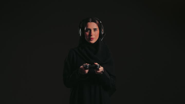 ايماءات الاندماج والتفاعل أثناء اللعب، استخدام التقنيات الالكترونية الحديثة والمتطورة ، شابة سعودية تلعب بألعاب الفيديو الترفيهية في عطلة نهاية الاسبوع، تصوير لإمرأة عربية خليجية إماراتية ترتدي العباءة والحجاب تحمل بيدها عصا التحكم