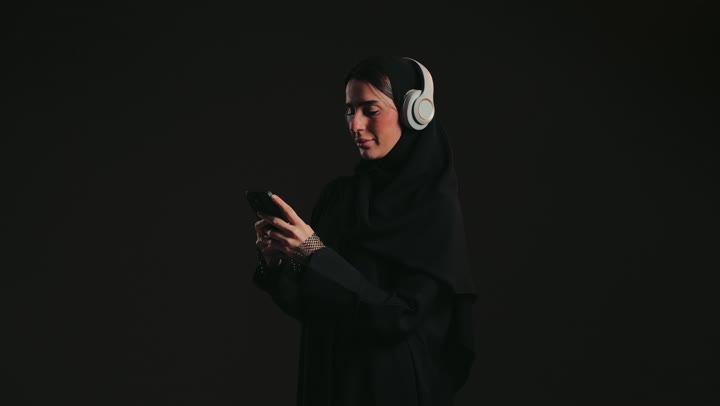 Having a good time listening to music, making a video call, using headphones in free time, photographing an Emirati Gulf Arab woman wearing an abaya and hijab, wearing headphones and holding a mobile phone, taking a souvenir selfie