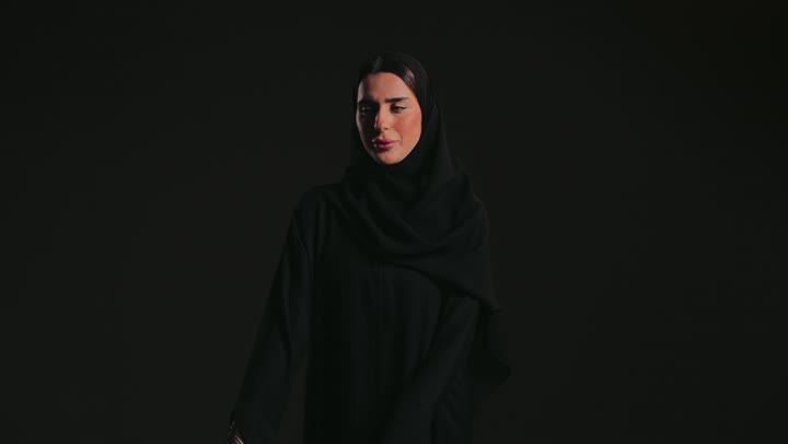 Facial and hand movements indicating deliberation and patience, a close-up of an Emirati Gulf Arab woman wearing a black abaya and hijab, raising her fist with gestures of deliberation and waiting, standing and looking at the camera with a cheerful expression.