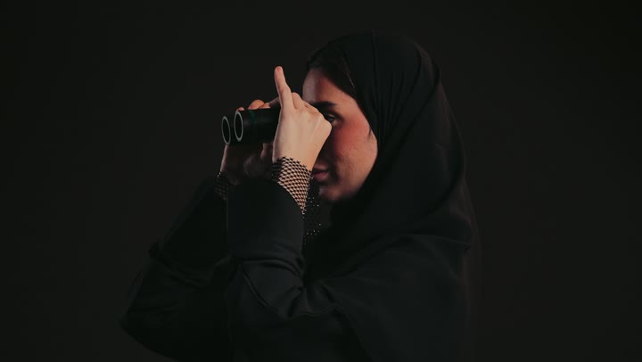 التفكير والتطلع إلى المستقبل، التخطيط المستقبلي للأهداف والطموحات، سيدة سعودية تقوم برؤية الأشياء البعيدة، حب الاستكشاف والمغامرة، تصوير لشابة عربية خليجية اماراتيه ترتدي العباءة السوداء والحجاب تمسك منظار ثنائي العينين تنظر إلى شيء ما .
