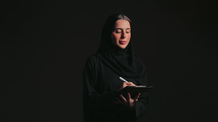 تنظيم وإدارة الأعمال, تدوين وتسجيل الملاحظات والمعلومات على الورق, تصوير لامرأة عربية خليجية اماراتية ترتدي العباءة السوداء والحجاب تقوم بكتابة المهام والأهداف اليومية .