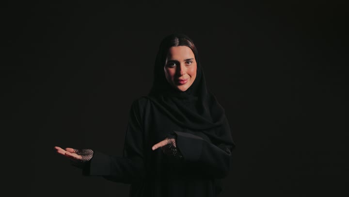 A Saudi woman locating something, taking care of her outward appearance, raising her hand with a gesture of identification, signs and hand movements indicating this place (here), a picture of an Emirati Gulf Arab woman wearing an abaya and hijab, pointing with her hands to the left.
