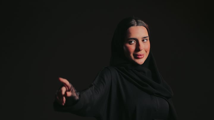 رفع اليد بايماءات التحديد ، إشارات وحركات يدين تدل على هذا المكان (هنا)، سيدة سعودية تقوم بتحديد موقع شيء معين، تصوير لامرأة عربية خليجية إماراتية ترتدي عباءة وحجاب تشير بيديها إلى جهة اليسار .