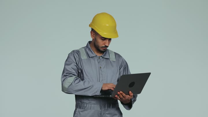 Integrating modern technologies into practical life, formal dress for work in the engineering sector, repair work and maintenance workshops in the UAE, an Arab Gulf Emirati technical engineer wearing a helmet and safety suit managing his work via laptop, youth professions and jobs, white background