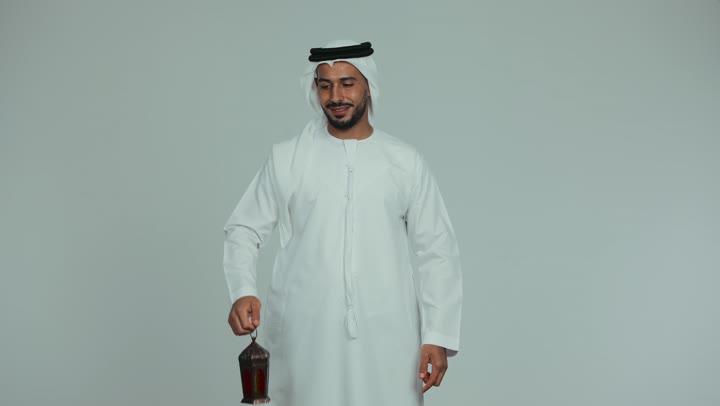 Preparing for the arrival of the holy month of Ramadan, looking at the camera with gestures of pleasure and happiness, an atmosphere of serenity and spirituality, celebrating religious occasions, an Arab Gulf Emirati man wearing a kandura and ghutra holding a metal lantern in his hand and pointing at it with his index finger, white background