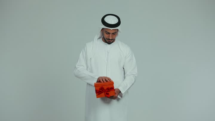 أعياد ومناسبات ومفاجآت سعيدة، رجل عربي خليجي إماراتي يرتدي الكندورة والغترة يحمل بيده علبة هدية يقوم بفتحها ينظر إلى داخلها بإيماءات الدهشة والسعادة، تقديم الهدايا بين الأهل والأصدقاء، التعبير عن المحبة والألفة، خلفية بيضاء
