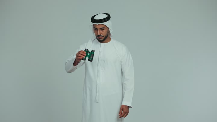 Seeing distant things, love of adventure and exploration, an Arab Gulf Emirati man wearing a kandura and ghutra holding a binocular (telescope), pointing his index finger at something, future planning for goals and ambitions, white background