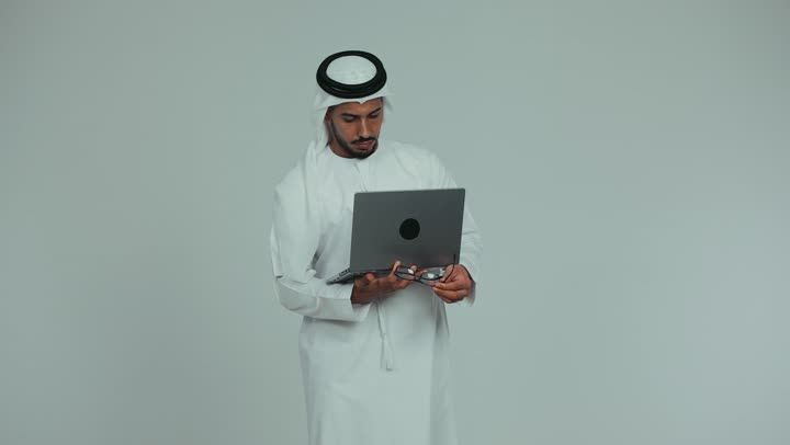 Managing and completing tasks using a modern and advanced technical device, an Emirati Gulf Arab man wearing a kandura, ghutra, and eyeglasses, working on a laptop with gestures and expressions of seriousness and focus, Emirati office jobs and professions, white background