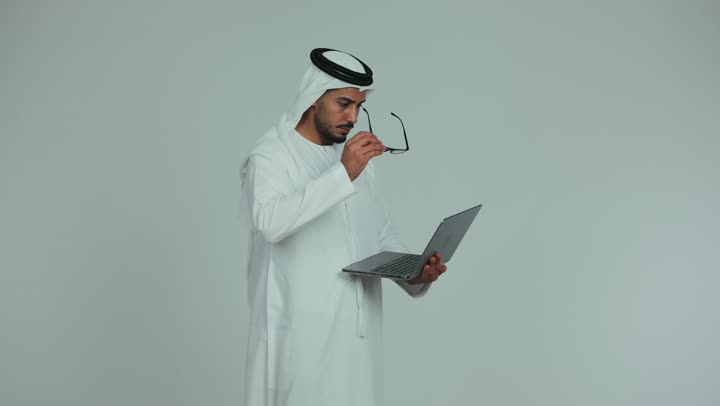 Managing and completing tasks using a modern and advanced technical device, an Emirati Gulf Arab man wearing a kandura, ghutra, and eyeglasses, working on a laptop with gestures and expressions of seriousness and focus, Emirati office jobs and professions, white background
