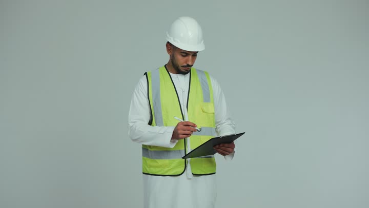 العمل في المجال الهندسي, مفهوم الهندسة و الإنشاءات المعمارية, الإشراف على الأعمال وتدقيقها، رجل عربي خليجي اماراتي يرتدي الكندورة والخوذة وسترة الحماية يقوم بتدوين الملاحظات على المخططات والمشاريع الهندسية، الإعمار بسواعد أبناء الوطن، خلفية بيضاء.