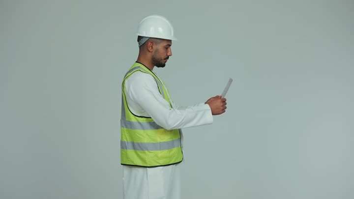 الإشراف على الأعمال وتدقيقها، العمل في المجال الهندسي, مفهوم الهندسة و الإنشاءات المعمارية, تصوير من الجانب لرجل عربي خليجي اماراتي يرتدي الكندورة والخوذة وسترة الحماية يقوم بتدقيق المخططات والمشاريع الهندسية، الإعمار بسواعد أبناء الوطن، خلفية بيضاء.