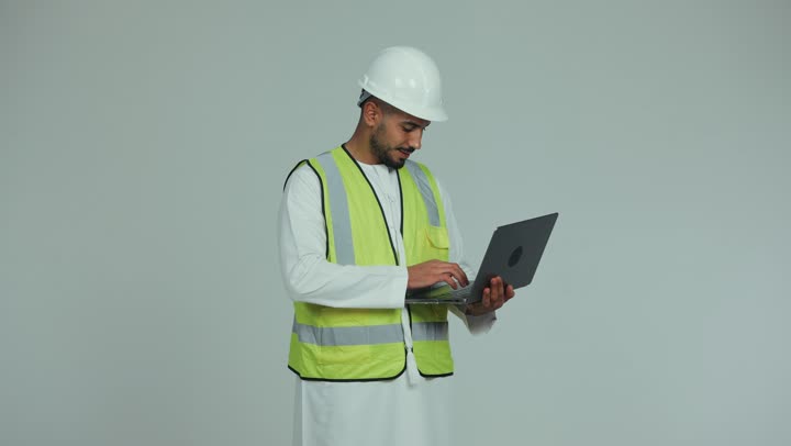 إدارة المشاريع باستخدام التقنيات الحديثة، بناء الوطن بسواعد أبنائه, رجل عربي خليجي اماراتي يرتدي الكندورة والخوذة وسترة الحماية يقوم بمتابعة وتدقيق المخططات الإنشائية باستخدام الحاسوب المحمول، العمل في القطاع الهندسي، خلفية بيضاء