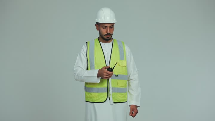 إعطاء أوامر خاصة بالعمل، مفهوم الهندسة و الإنشاءات, مهندس عربي خليجي اماراتي رتدي الخوذة وسترة الحماية يقوم بالتحدث عبر الجهاز اللاسلكي  للإشراف على الأعمال ومتابعتها، المخططات والمشاريع الهندسية، خلفية بيضاء.