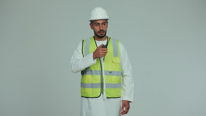 مفهوم الهندسة و الإنشاءات, الإشارة بإصبع السبابة إلى شيء ما، إعطاء أوامر خاصة بالعمل، مهندس عربي خليجي اماراتي رتدي الخوذة وسترة الحماية يقوم بالتحدث عبر الجهاز اللاسلكي  للإشراف على الأعمال ومتابعتها، المخططات والمشاريع الهندسية، خلفية بيضاء.
